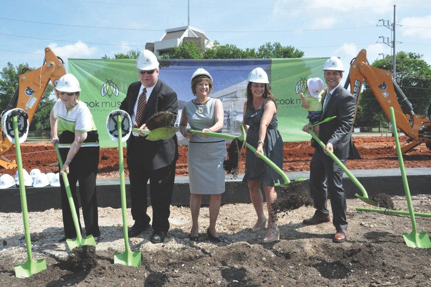 Five individuals in hard hats and formal attire are performing a ceremonial groundbreaking with shovels, standing on a construction site with a banner for the Cook Museum in the background.