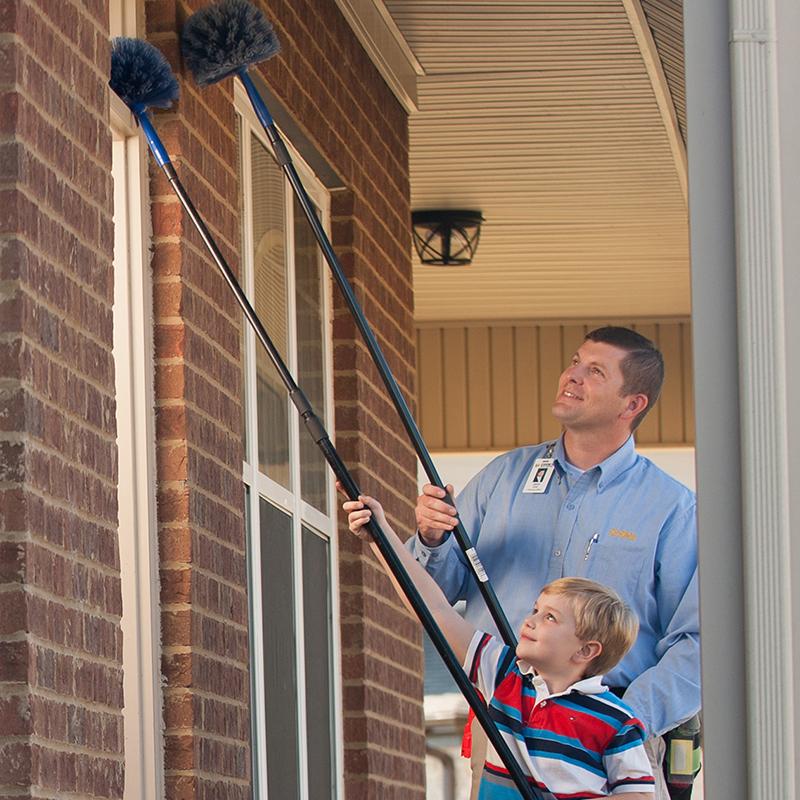 A Cook's pest control professional and a young boy using cobweb dusters on a house exterior.