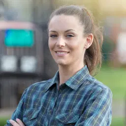 headshot of Young pretty farmer woman standing on farmland with crossed arms and tractors in background