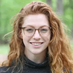 headshot of Young woman with glasses smiling at the camera with an outdoors background.