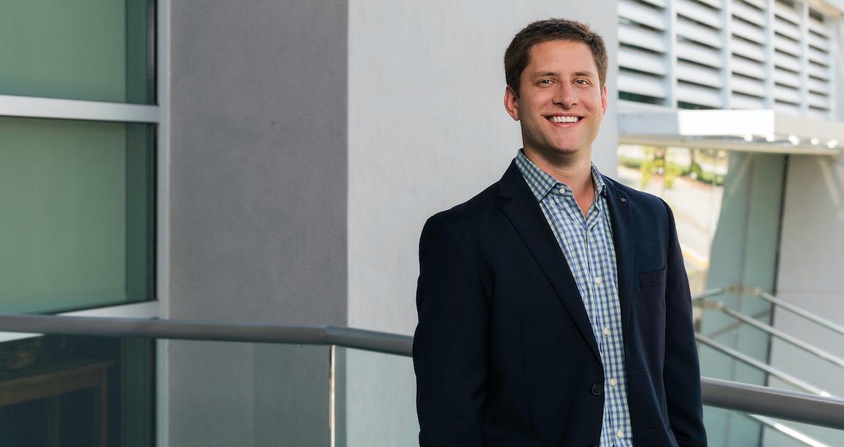 Man in suit smiling on a balcony with a modern building backdrop.