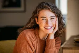 headshot of Joyful woman with curly hair in a rust sweater, leaning on hand.