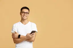 headshot of Young man in glasses and white tee with phone, arms crossed on beige background.