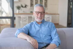 headshot of Smiling older man with glasses sitting on a couch indoors.