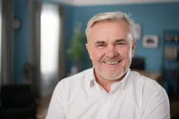 headshot of Smiling older man in white shirt smiling indoors with a blue room in the background.